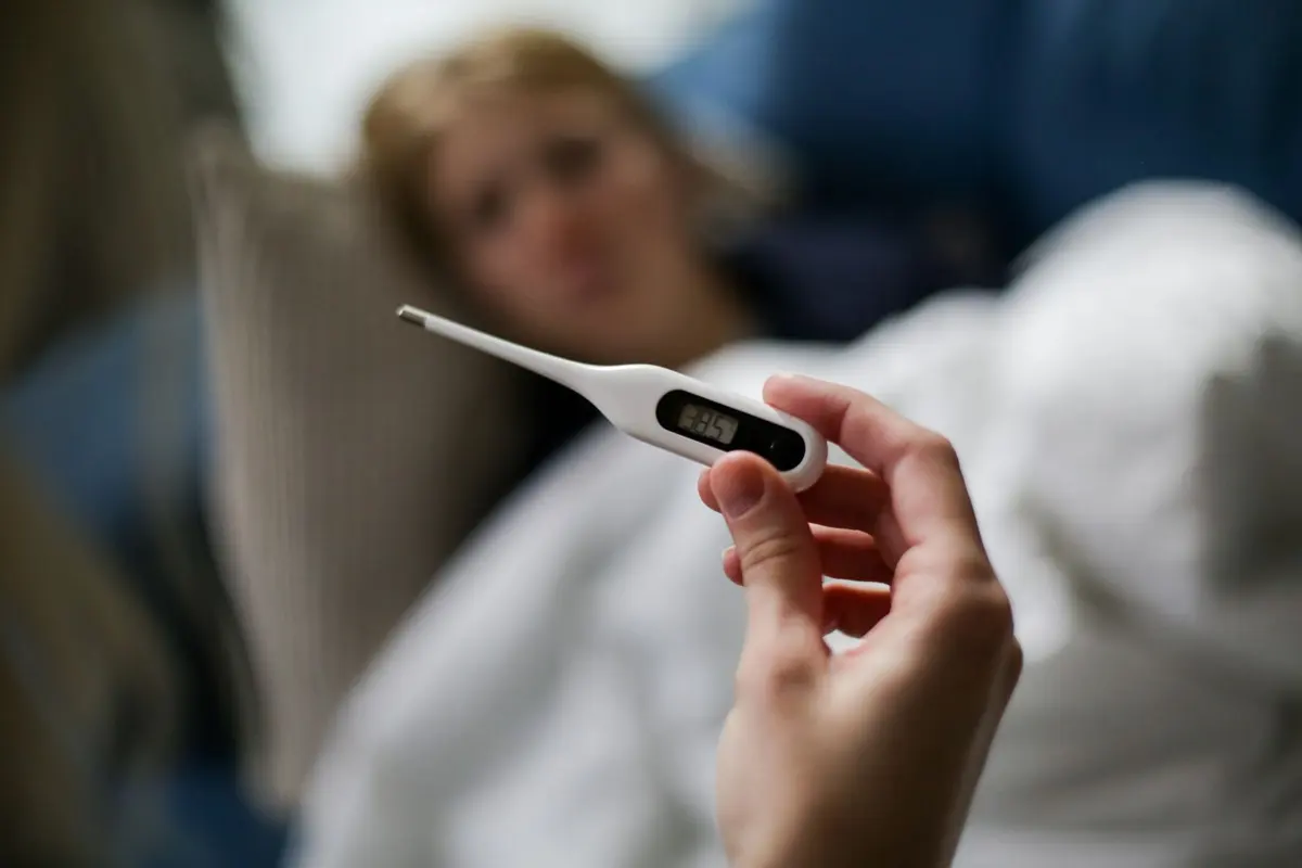 Hand holding a digital thermometer while a person with fever rests in bed, tracking dengue symptoms day by day