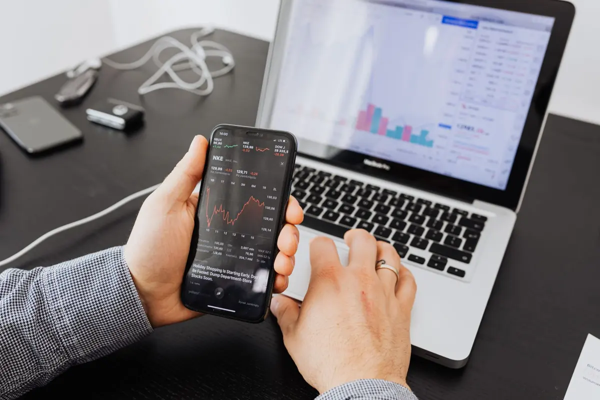 Person checking a market price chart on a smartphone in front of a laptop