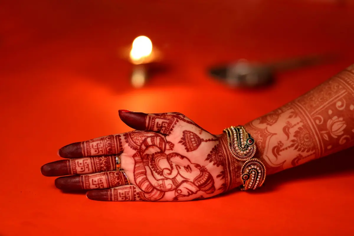 A hand decorated with detailed bridal henna next to a lit diya on an orange background, ready for Karwa Chauth