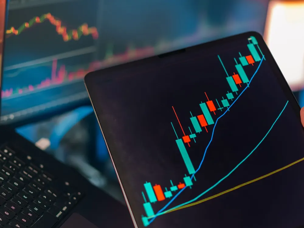 Tablet showing a rising candlestick stock chart in front of a laptop with market data