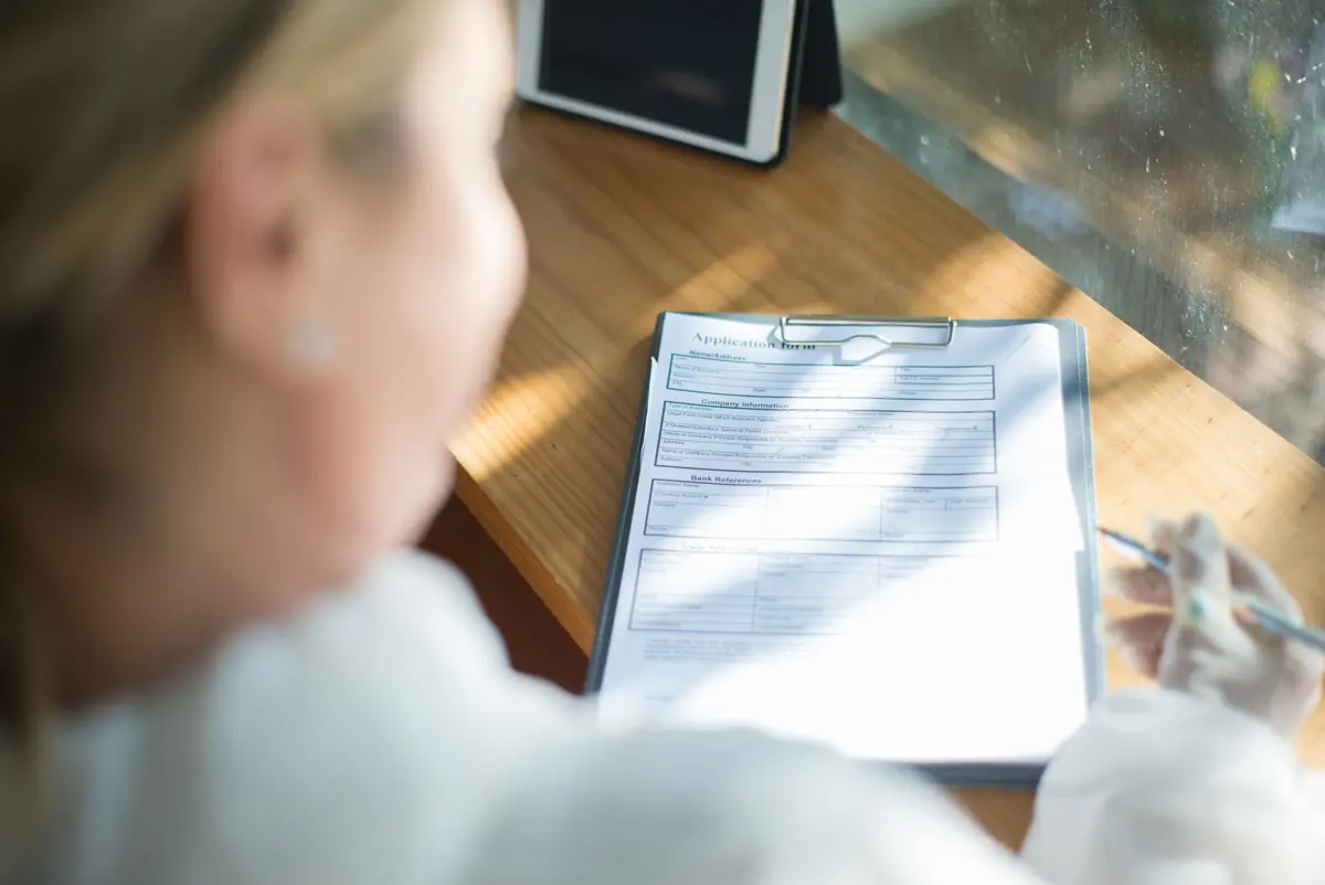 A woman filling out a paper application form on a clipboard at a sunlit wooden desk